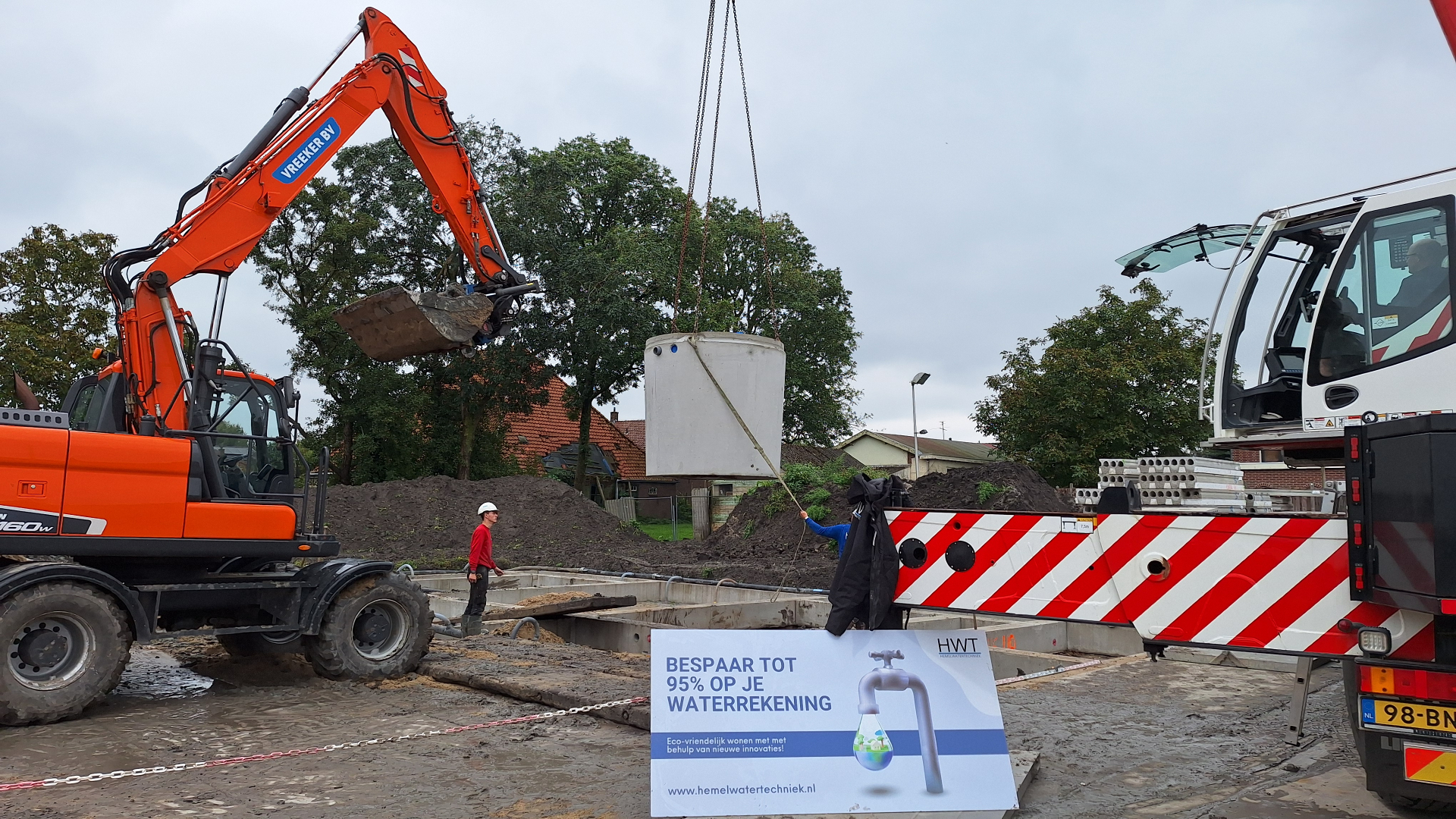 Een kraandrijver een betonnen regenwatertank met grote precisie zakken binnen de fundering van een van elf woningen in Venhuizen - foto: Margo van Voskuilen
